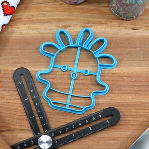 May include: A blue plastic cookie cutter shaped like a cartoon character with a large head and a small body. The character has two large eyes and a small mouth. The cookie cutter is on a wooden surface.