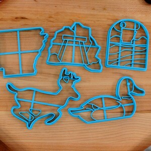Op de afbeelding: Vijf blauwe plastic koekjesvormen op een houten oppervlak. De vormen zijn in de vorm van een hert, een eend, een raam, een brug en een staatsschets.