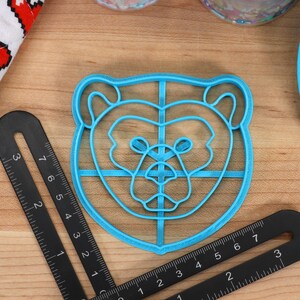 May include: A blue plastic bear-shaped cookie cutter with a grid pattern inside. The cutter is on a wooden surface.