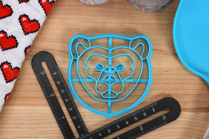 May include: A blue plastic bear-shaped cookie cutter with a grid pattern inside. The cutter is on a wooden surface.