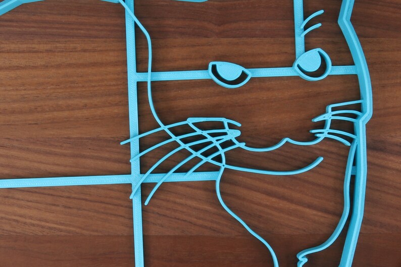 May include: A blue 3D printed cat outline on a wooden surface. The cat's face is visible with eyes, whiskers, and a smile.