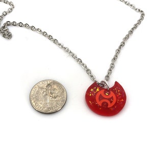 May include: A red and orange glitter necklace with a silver chain and a pendant shaped like a crescent moon with a stylized symbol inside. The pendant is about the size of a US dime.