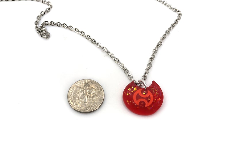 May include: A red and orange glitter necklace with a silver chain and a pendant shaped like a crescent moon with a stylized symbol inside. The pendant is about the size of a US dime.
