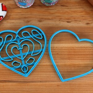 May include: Two blue heart-shaped cookie cutters, one with a detailed design and the other plain, on a wooden surface.