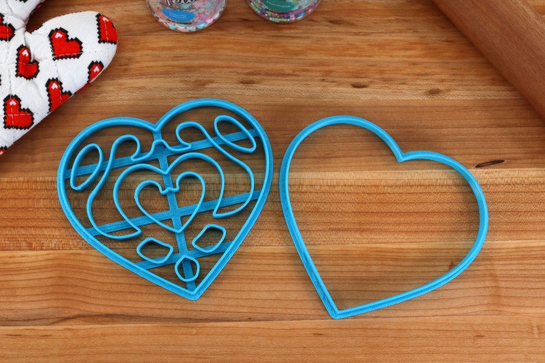 May include: Two blue heart-shaped cookie cutters, one with a detailed design and the other plain, on a wooden surface.
