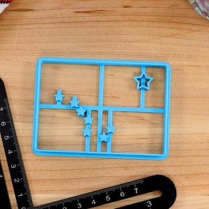 May include: A blue plastic cookie cutter shaped like a grid with nine star-shaped cutouts. The cookie cutter is on a wooden surface.