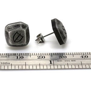 May include: A pair of silver stud earrings with a textured, geometric design. Each earring features a raised, shield-shaped symbol. The earrings are shown next to a ruler, measuring approximately 1 cm in diameter.