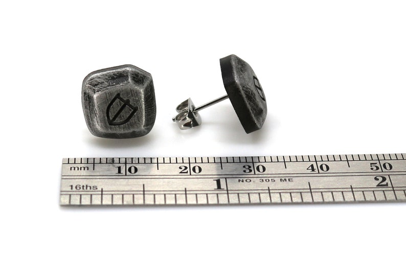 May include: A pair of silver stud earrings with a textured, geometric design. Each earring features a raised, shield-shaped symbol. The earrings are shown next to a ruler, measuring approximately 1 cm in diameter.