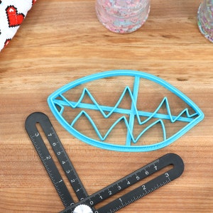 May include: A blue plastic cookie cutter shaped like an eye with a wavy design inside. The cutter is on a wooden surface.