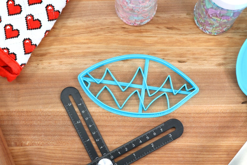 May include: A blue plastic cookie cutter shaped like an eye with a wavy design inside. The cutter is on a wooden surface.