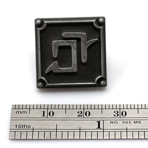 K&ouml;nnte beinhalten: Quadratischer, antik-silberfarbener Metallstift mit einem abstrakten Design. Der Stift hat einen erhabenen Rand mit abgerundeten Ecken und ein stilisiertes Symbol in der Mitte. Ein Lineal ist darunter platziert, mit Messungen in Zentimetern.