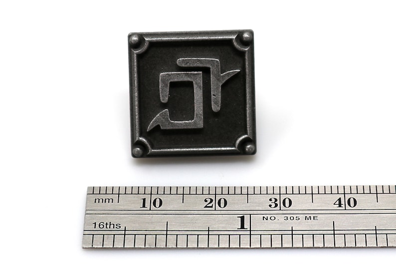 K&ouml;nnte beinhalten: Quadratischer, antik-silberfarbener Metallstift mit einem abstrakten Design. Der Stift hat einen erhabenen Rand mit abgerundeten Ecken und ein stilisiertes Symbol in der Mitte. Ein Lineal ist darunter platziert, mit Messungen in Zentimetern.