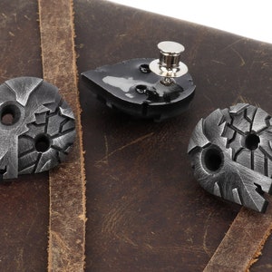 May include: Three silver metal guitar picks with a textured, distressed finish. The picks are shaped like a teardrop and have two holes each. One pick is on a brown leather surface.