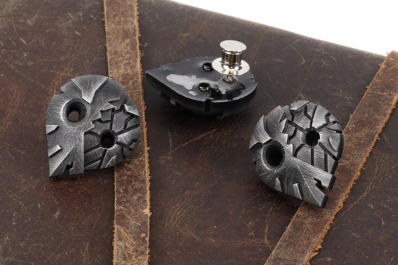 May include: Three silver metal guitar picks with a textured, distressed finish. The picks are shaped like a teardrop and have two holes each. One pick is on a brown leather surface.