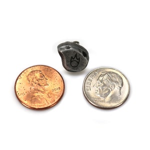 May include: A small, irregularly shaped, dark gray metal pendant with a flame symbol, next to a penny and a dime. The pendant is approximately 0.75 inches in length. The items are on a white background.