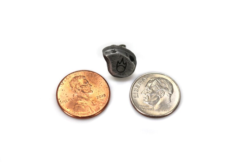 May include: A small, irregularly shaped, dark gray metal pendant with a flame symbol, next to a penny and a dime. The pendant is approximately 0.75 inches in length. The items are on a white background.