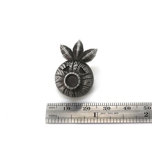 以下が含まれることがあります： 丸い体と上に3枚の葉を持つ顔の形をした銀色の金属製のペンダント。顔は黒い目を2つと大きく開いた口をしています。ペンダントの直径は約2cmです。