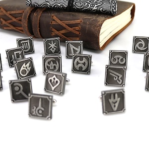 May include: A set of 18 silver cufflinks with different symbols, including a sword, a shield, a fire, and a star. The cufflinks are arranged on a white surface.