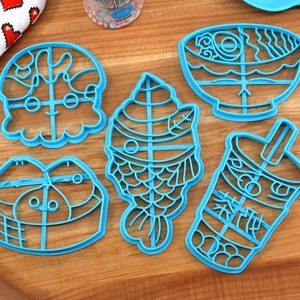 May include: Six blue plastic cookie cutters in various shapes, including a fish, a bowl of ramen, a bubble tea, and a fish-shaped ice cream cone. The cutters are on a wooden surface with a rolling pin and a heart-patterned cloth.