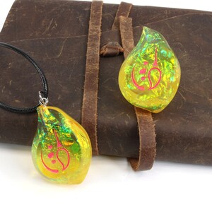 May include: Two iridescent yellow teardrop-shaped pendants with a red symbol inside. One pendant is on a black cord necklace, and the other is lying on a brown leather journal.