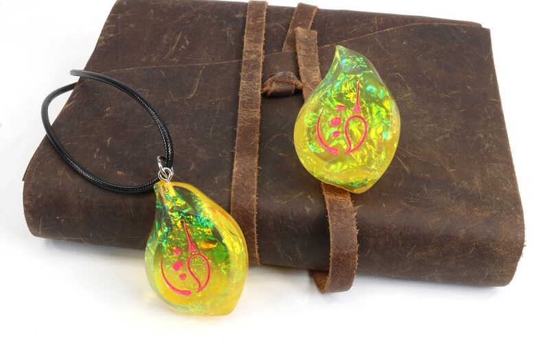 May include: Two iridescent yellow teardrop-shaped pendants with a red symbol inside. One pendant is on a black cord necklace, and the other is lying on a brown leather journal.