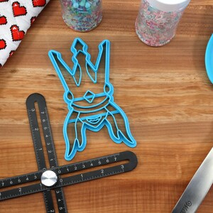 May include: A light blue cookie cutter in the shape of a crown sits on a wooden surface, next to a blue spatula, a black angle ruler, and a heart-patterned oven mitt. Sprinkles are in the background.