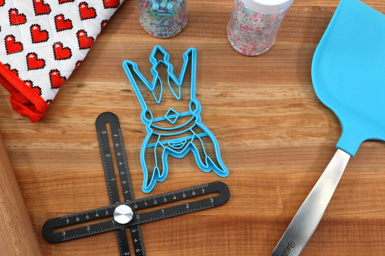 May include: A light blue cookie cutter in the shape of a crown sits on a wooden surface, next to a blue spatula, a black angle ruler, and a heart-patterned oven mitt. Sprinkles are in the background.