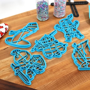 May include: A set of five blue plastic cookie cutters shaped like knights and a ship. The cutters are on a wooden surface.