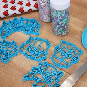 May include: Five blue plastic cookie cutters shaped like cartoon characters, a container of colorful sprinkles, and a blue spatula on a wooden surface.
