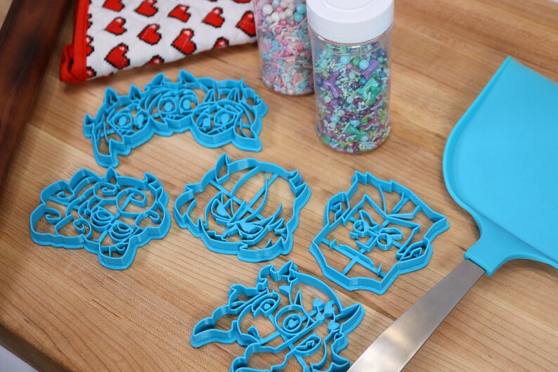 May include: Five blue plastic cookie cutters shaped like cartoon characters, a container of colorful sprinkles, and a blue spatula on a wooden surface.
