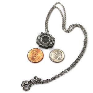 Puede incluir: Un collar de plata con un colgante con forma de dispositivo de videojuego. El colgante es del tama&ntilde;o de una moneda de un centavo de d&oacute;lar estadounidense. El collar est&aacute; sobre un fondo blanco.