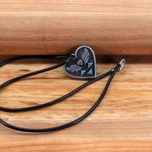 Puede incluir: Un collar de cord&oacute;n negro con un colgante de coraz&oacute;n plateado con un s&iacute;mbolo alado. El colgante est&aacute; sobre una superficie de madera.