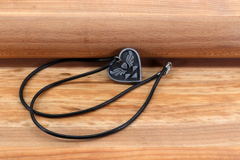 Puede incluir: Un collar de cord&oacute;n negro con un colgante de coraz&oacute;n plateado con un s&iacute;mbolo alado. El colgante est&aacute; sobre una superficie de madera.