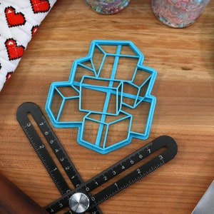 May include: A blue plastic cookie cutter shaped like a stack of cubes. The cutter is on a wooden surface with a ruler, a rolling pin, and a spatula.