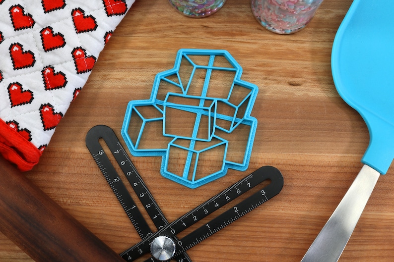 May include: A blue plastic cookie cutter shaped like a stack of cubes. The cutter is on a wooden surface with a ruler, a rolling pin, and a spatula.