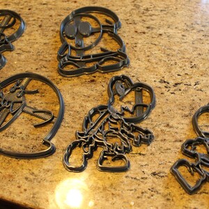May include: Five gray plastic cookie cutters shaped like cartoon characters. The cutters are on a brown speckled countertop.