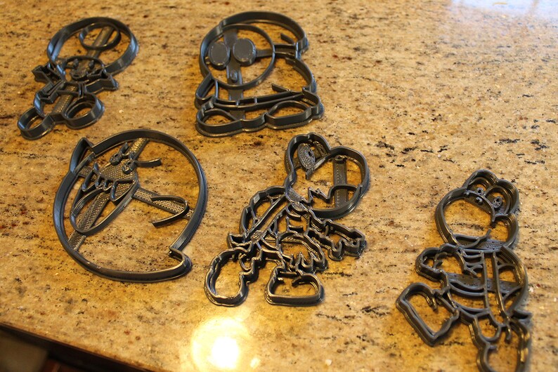 May include: Five gray plastic cookie cutters shaped like cartoon characters. The cutters are on a brown speckled countertop.