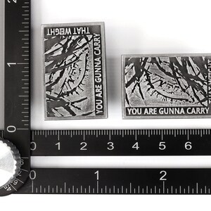 May include: Two silver metal pins with a distressed, textured surface. The pins feature the text "You are gunna carry that weight" in black lettering.