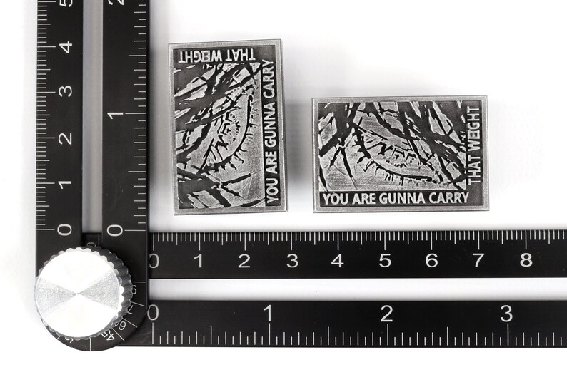 May include: Two silver metal pins with a distressed, textured surface. The pins feature the text "You are gunna carry that weight" in black lettering.