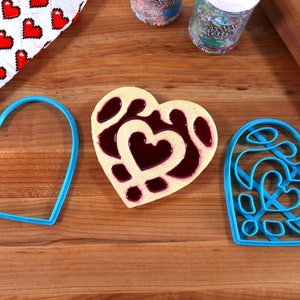 May include: Three blue heart-shaped cookie cutters on a wooden surface. One cookie cutter is filled with a heart-shaped cookie with a red jelly filling.