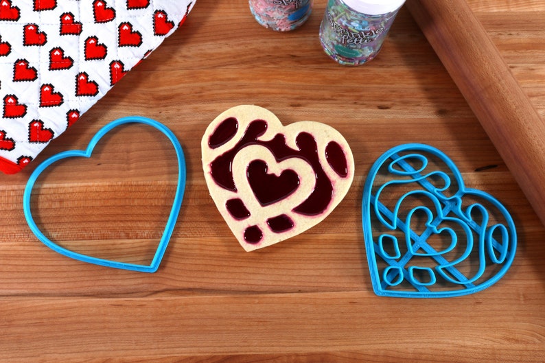 May include: Three blue heart-shaped cookie cutters on a wooden surface. One cookie cutter is filled with a heart-shaped cookie with a red jelly filling.