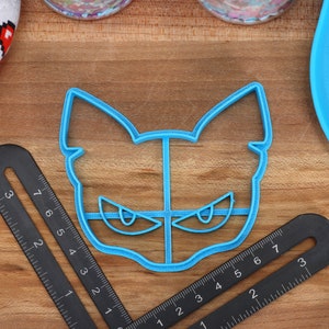 May include: A blue plastic cookie cutter shaped like a cat's face with angry eyes. The cutter is on a wooden surface with a ruler.