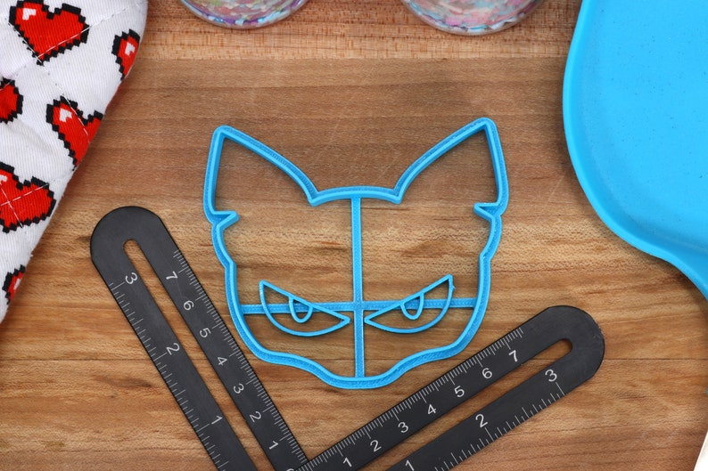 May include: A blue plastic cookie cutter shaped like a cat's face with angry eyes. The cutter is on a wooden surface with a ruler.
