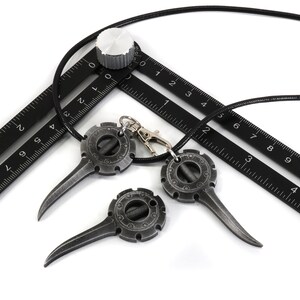 May include: Three black metal pendants with a circular design and a pointed tip hanging from a black cord. The pendants are attached to a black ruler with white markings.