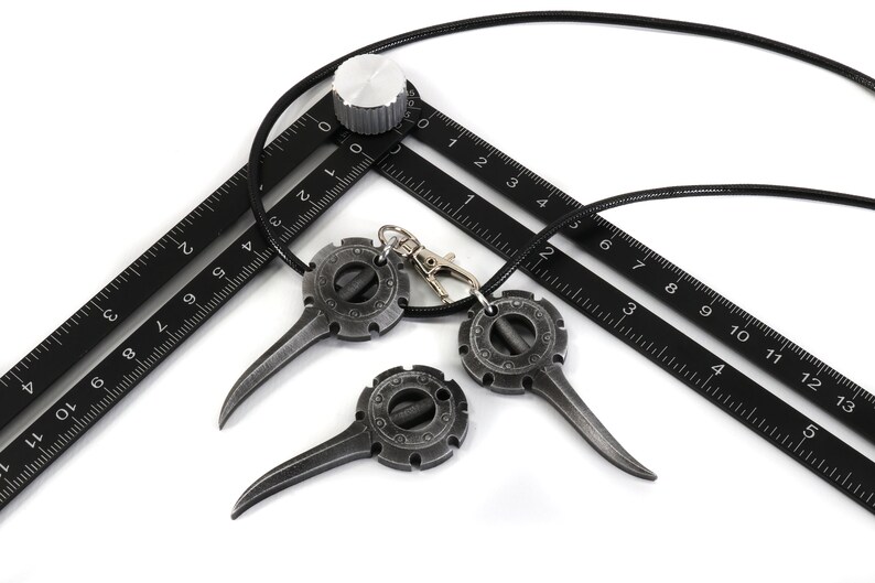 May include: Three black metal pendants with a circular design and a pointed tip hanging from a black cord. The pendants are attached to a black ruler with white markings.