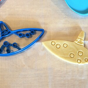 May include: A blue plastic cookie cutter shaped like an ocarina, next to a light brown cookie cut with the same shape.