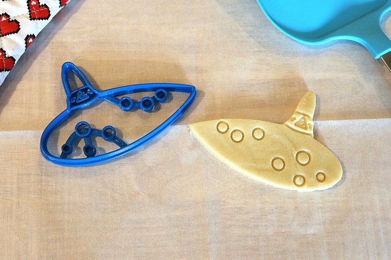 May include: A blue plastic cookie cutter shaped like an ocarina, next to a light brown cookie cut with the same shape.