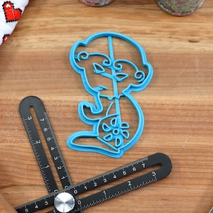 May include: A blue plastic cookie cutter shaped like a cartoon monkey holding a lollipop. The cookie cutter is on a wooden surface.