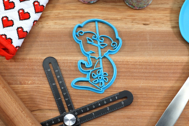 May include: A blue plastic cookie cutter shaped like a cartoon monkey holding a lollipop. The cookie cutter is on a wooden surface.