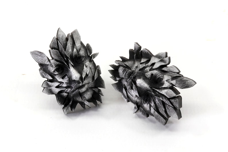 Pu&ograve; includere: Un paio di orecchini in metallo color argento a forma di viso con un design geometrico.